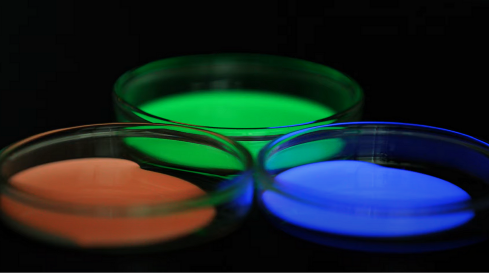 Das Bild zeigt drei Petrischalen, die Substanzen in verschiedenen Farben enthalten: Eine leuchtet grün, eine andere blau und die dritte orange. Dabei handelt es sich um BrightSplash™, druckbare Röntgenszintillatoren von BrightComSol, in Farben, die für verschiedene Detektoranwendungen optimiert sind. Die Schalen sind auf einer dunklen Oberfläche platziert, und der Hintergrund ist ebenfalls dunkel, was die Leuchtkraft der Farben verstärkt. © BrightComSol